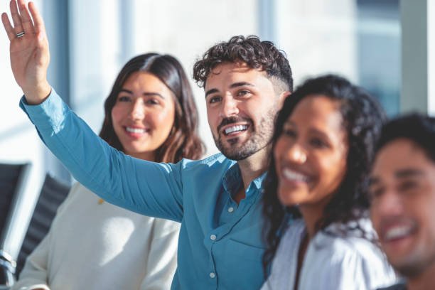 Group of people listening to a presentation. One person has their hand raised. They are at a seminar or meeting sitting at a table. They are all looking to the front of the room. Men and women dressed in casual business attire. Multi-cultural group with African, Latino and Caucasian